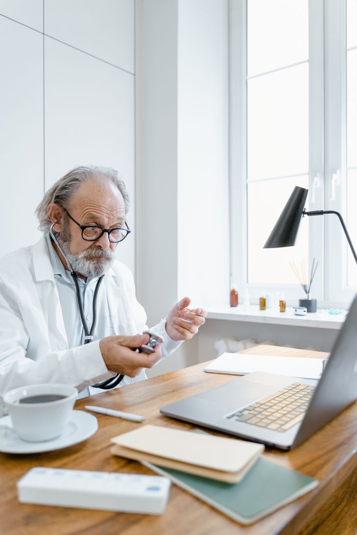 Senior doctor holding stethoscope during online consultation in office environment.