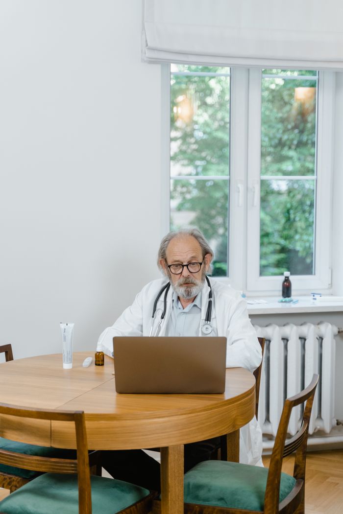 Senior doctor in office conducting video call on laptop, wearing eyeglasses and stethoscope.