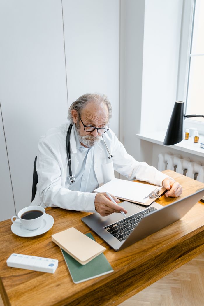 A senior doctor engaging in an online consultation with a laptop, notepad, and coffee cup.