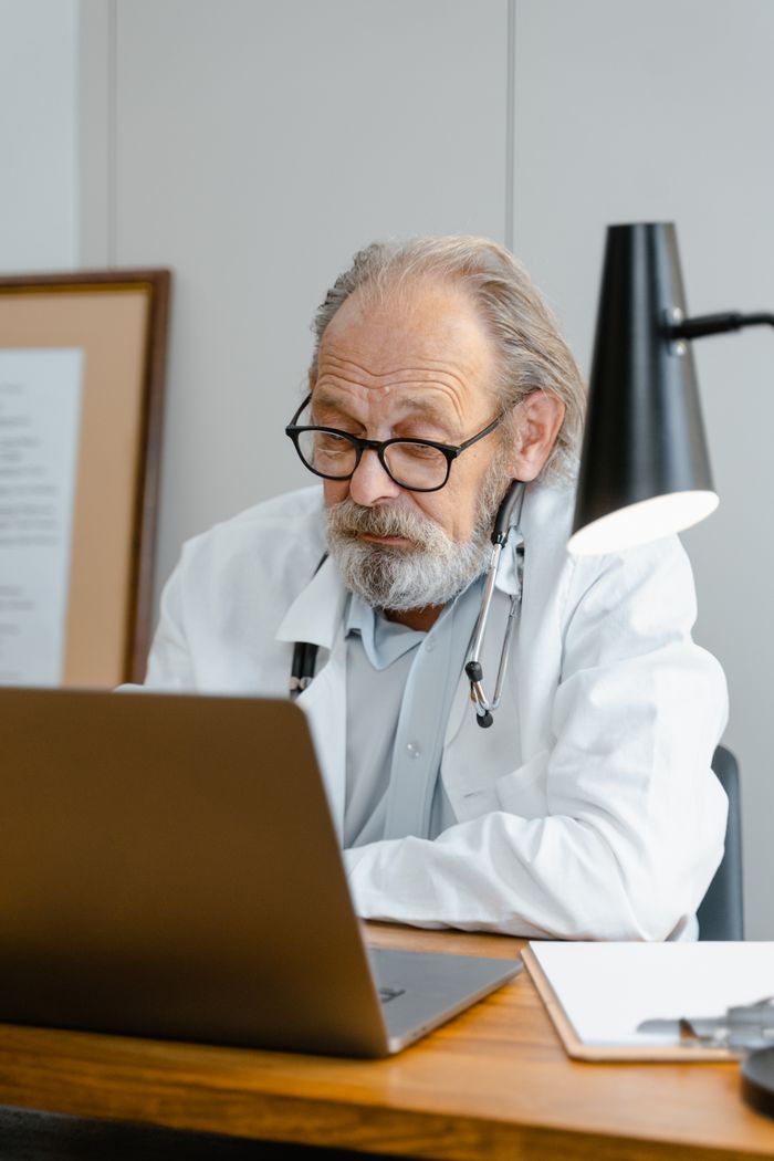 Senior male doctor reviewing patient records on his laptop in a cozy office setting.